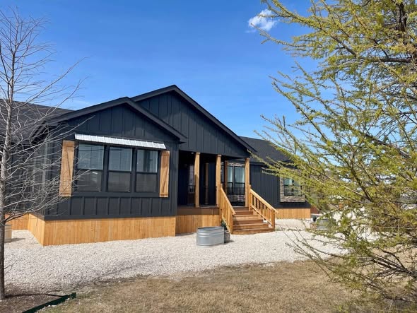 A modern black house with wooden accents and stairs sits on a gravel path, surrounded by sparse trees and grass under a clear blue sky.
