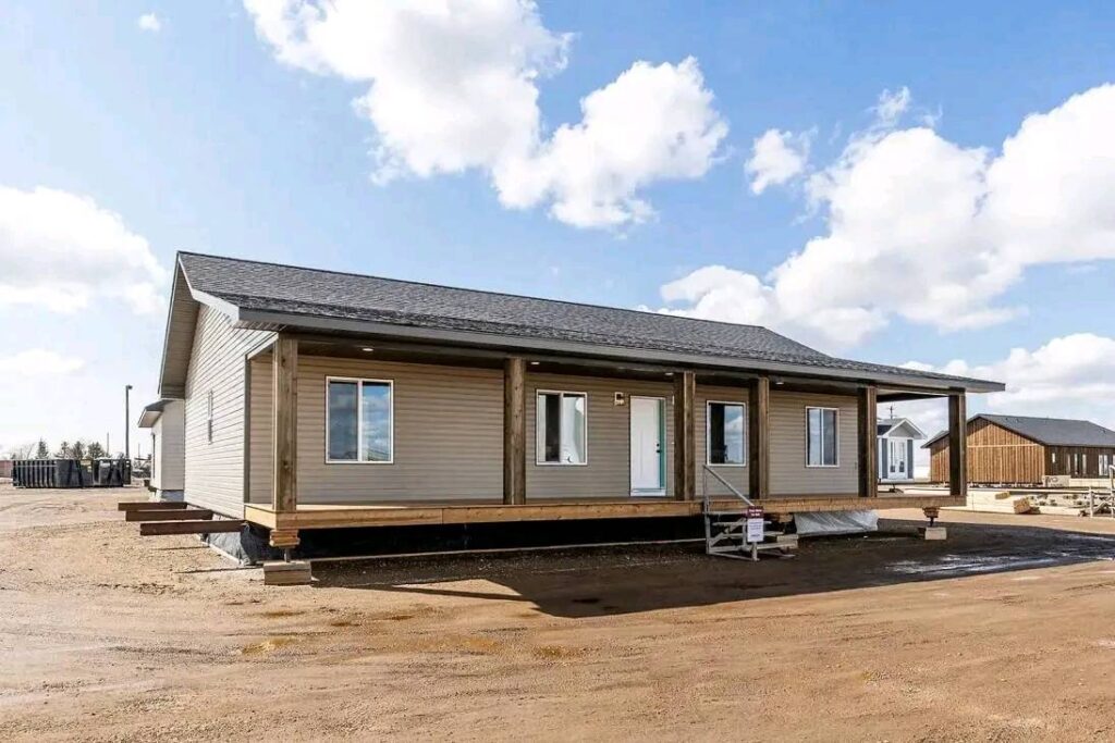 Single-story modular home with beige siding and a dark roof stands on a dirt lot under a blue sky with scattered clouds, conveying simplicity and spaciousness.