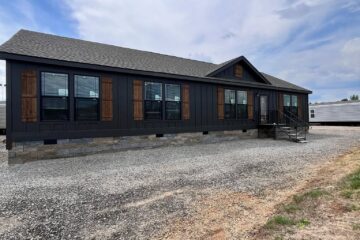 A modern, dark-colored prefab home with large windows and wooden shutters sits on a gravel lot. The sky is partly cloudy, creating a serene atmosphere.