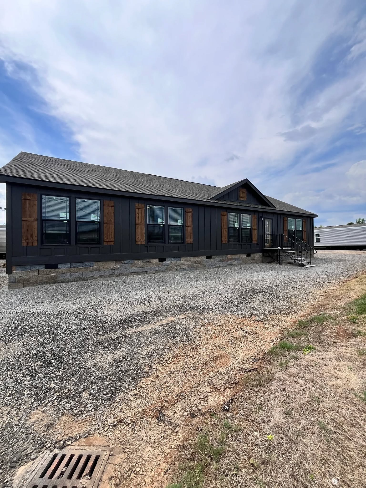A modern, dark-colored prefab home with large windows and wooden shutters sits on a gravel lot. The sky is partly cloudy, creating a serene atmosphere.