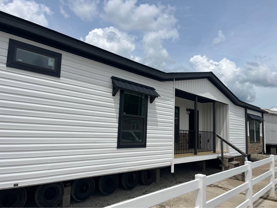 A white manufactured home with black trim, resting on wheels. It has a covered porch and a railing, set against a cloudy sky. The mood feels calm.