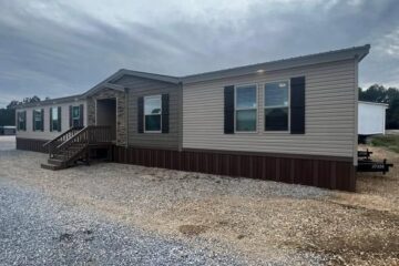 A beige manufactured home with dark shutters and a small porch sits on a gravel lot under a cloudy sky. The mood is calm and neutral.