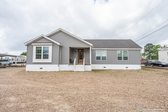 A single-story, gray manufactured home with a white-trimmed exterior sits on a gravel lot. The sky is cloudy, adding a calm, serene atmosphere.