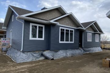 Single-story house with dark gray siding, white window frames, and gabled roof. Plastic covers the foundation. "For Sale" sign visible. Cloudy sky.