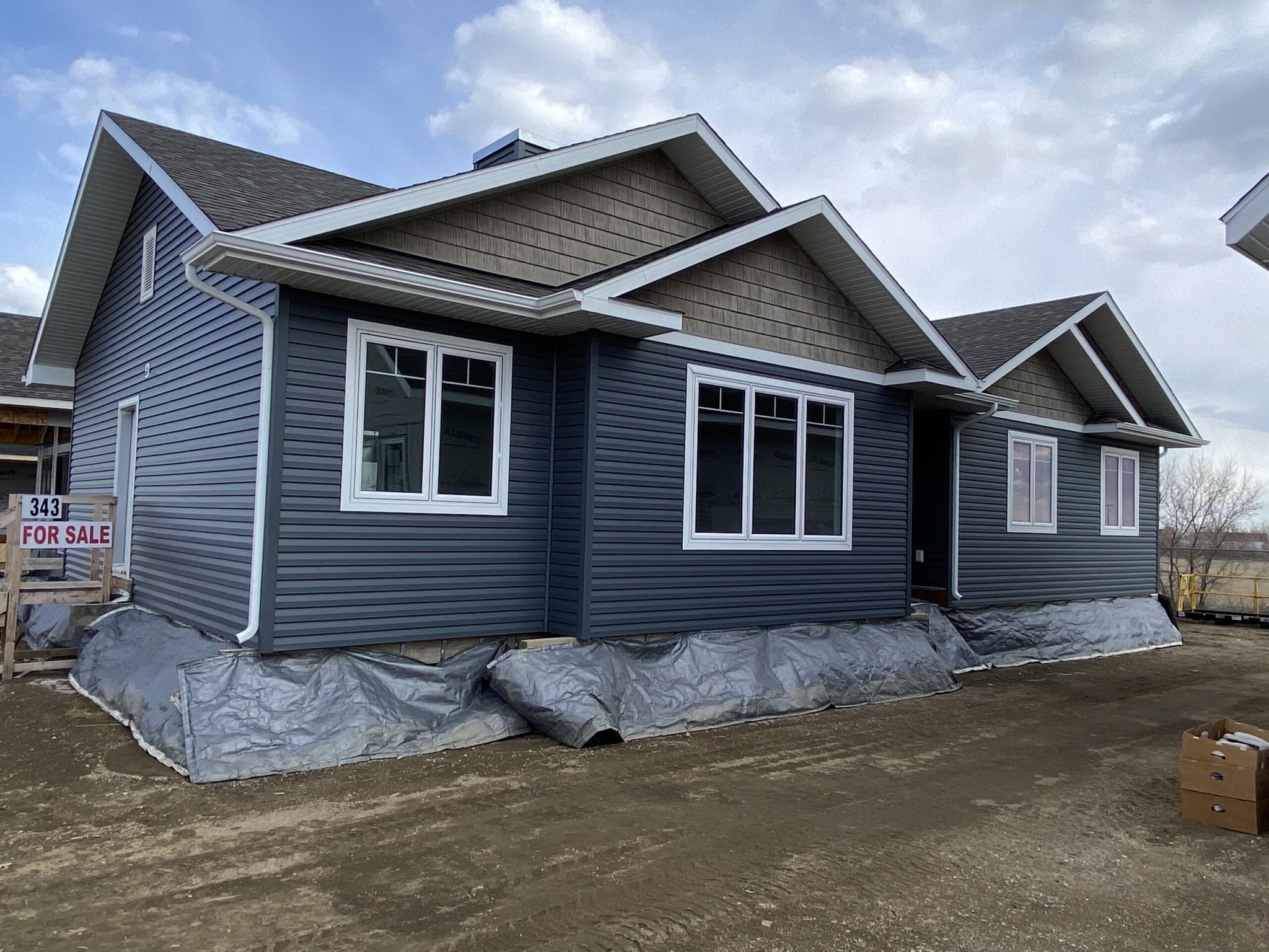 Single-story house with dark gray siding, white window frames, and gabled roof. Plastic covers the foundation. "For Sale" sign visible. Cloudy sky.