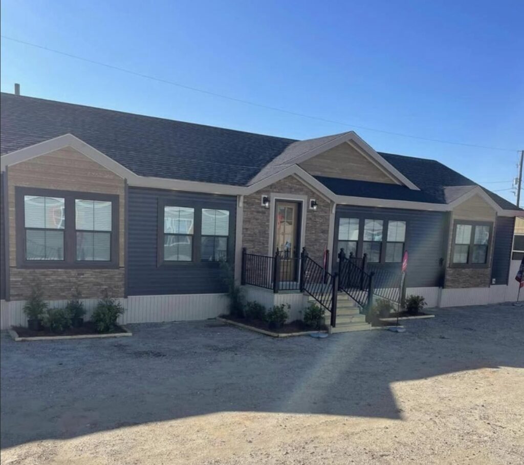 A modern modular home with a stone and siding exterior under a clear blue sky. Front porch features railings and small shrubs line the entrance.