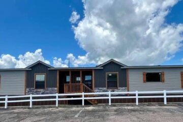 A modern manufactured home with a gray facade and brown shutters under a vibrant blue sky with fluffy clouds. A white fence surrounds the home, conveying a welcoming and serene atmosphere.
