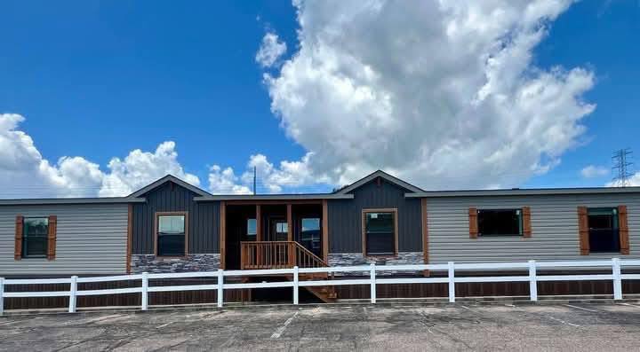 A modern manufactured home with a gray facade and brown shutters under a vibrant blue sky with fluffy clouds. A white fence surrounds the home, conveying a welcoming and serene atmosphere.