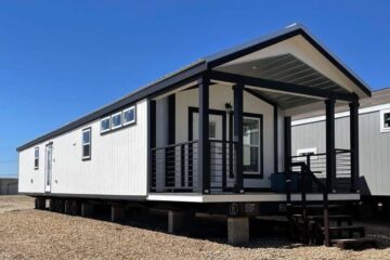 A modern, white mobile home with black trim sits elevated on a gravel lot under a clear blue sky. It features a small porch with steps and railing.