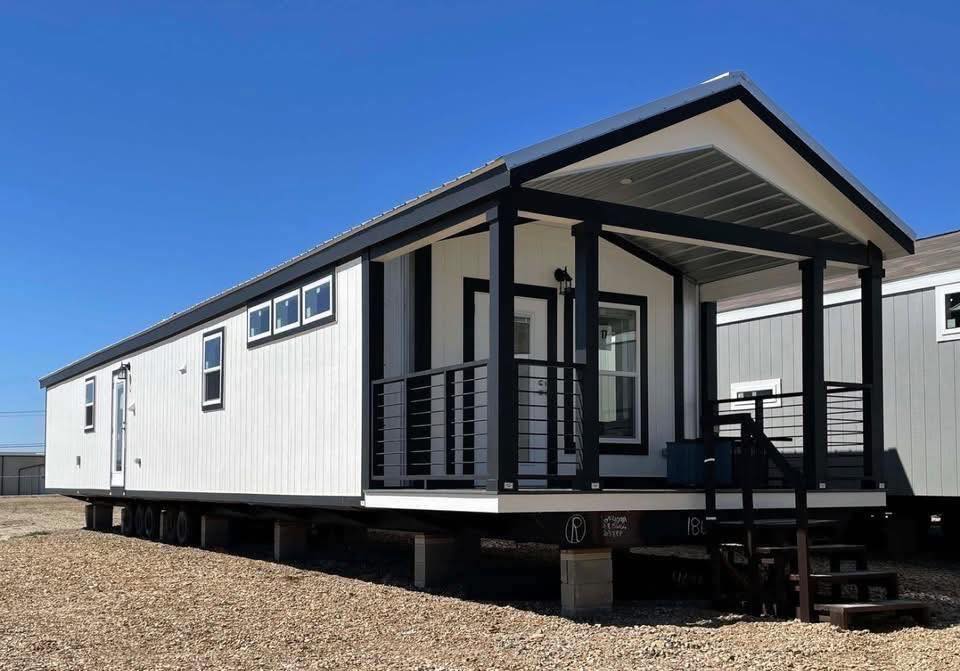 A modern, white mobile home with black trim sits elevated on a gravel lot under a clear blue sky. It features a small porch with steps and railing.
