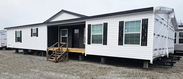 A white manufactured home with black shutters is set on a gravel lot. It features a small wooden porch with stairs leading to a black door. The tone is neutral.