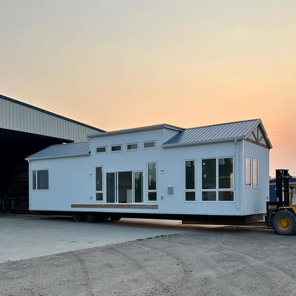 Modern white mobile home on wheels outside a warehouse, with large windows and a gabled roof. A forklift is positioned beside it at sunset.