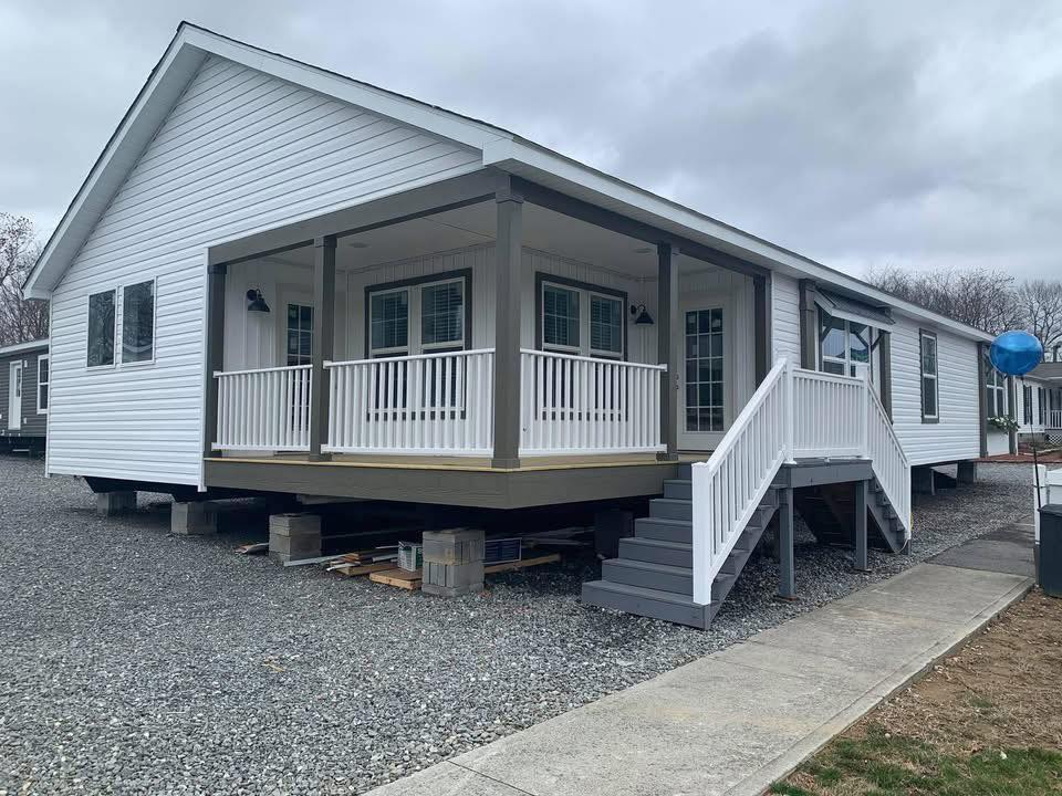 White modular home with a covered porch and steps, set on a gravel lot under a cloudy sky. The home has a modern, inviting appearance.