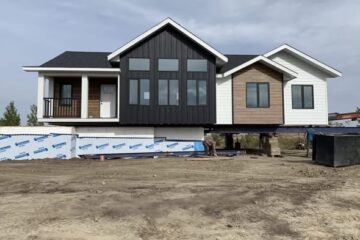 A modern house is elevated on piers with unfinished ground below. It features black, wood, and white siding, large windows, and a covered porch. Overcast sky above.