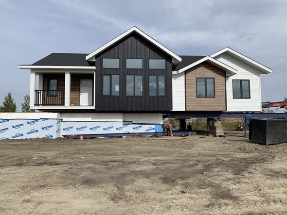 A modern house is elevated on piers with unfinished ground below. It features black, wood, and white siding, large windows, and a covered porch. Overcast sky above.
