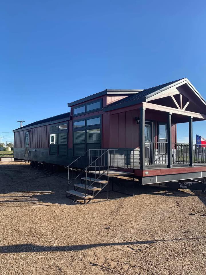 Rustic tiny house with red siding and black trim, surrounded by gravel. Steps lead to a porch. The clear blue sky adds a serene feel to the scene.