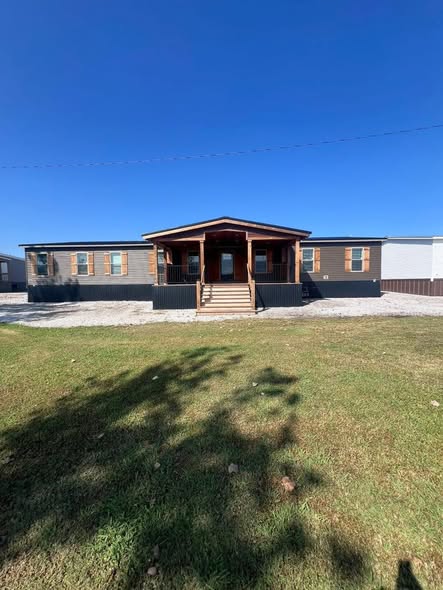 A modern manufactured home with a wooden entrance and steps sits under a clear blue sky. The front yard is grassy, casting tree shadows on the lawn.