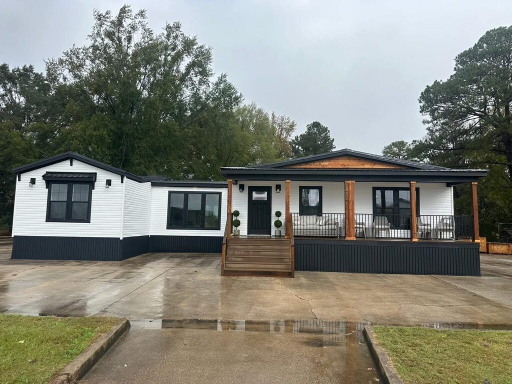 Single-story modern house with white siding, dark trim, and wooden columns. It features a large porch and is surrounded by lush green trees on a cloudy day.