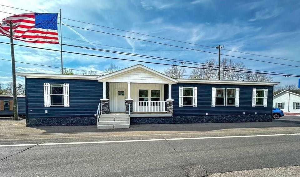 Single-story navy blue modular home with white shutters and a porch, under a bright blue sky. A large American flag flies prominently to the left.
