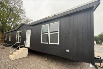 Dark-colored tiny house with a pitched roof and white door, set on gravel. Features include large windows and simple steps, surrounded by trees and a cloudy sky.