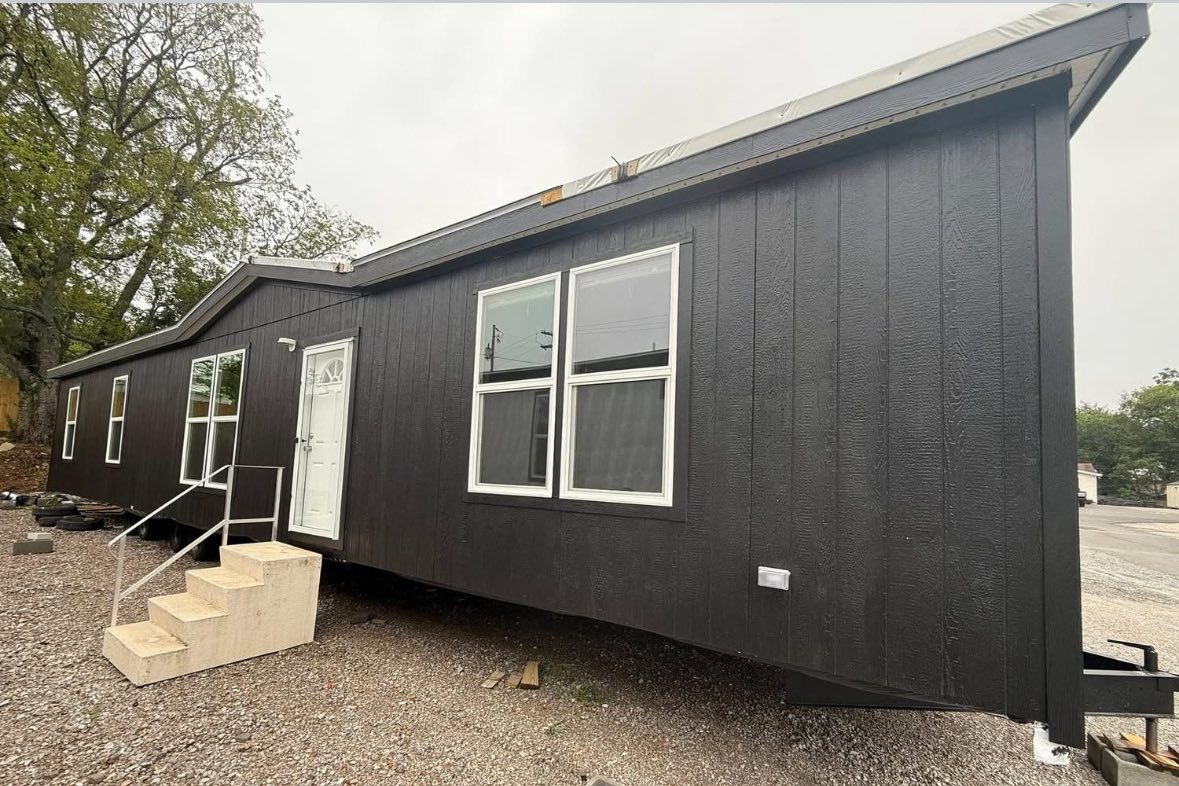 Dark-colored tiny house with a pitched roof and white door, set on gravel. Features include large windows and simple steps, surrounded by trees and a cloudy sky.