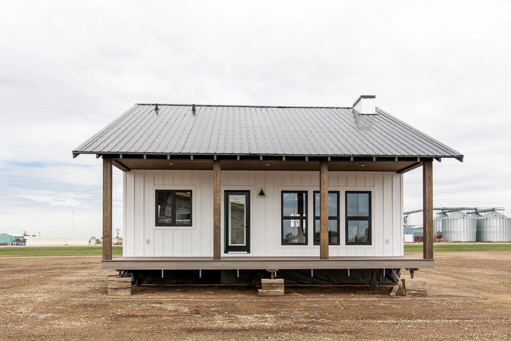 A small, modern white house with a metal roof and wooden pillars stands on an open field. Large windows and a central door convey a minimalist style.