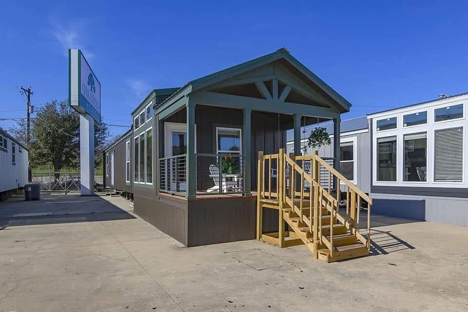 A modern tiny house with wooden steps and a small porch is showcased outdoors. It's flanked by similar structures, under a clear blue sky.