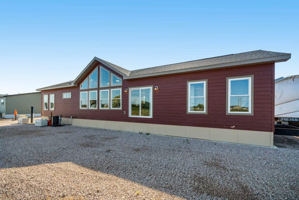A red, single-story modular home with a peaked roof and multiple large windows stands on a gravel lot under a clear blue sky, conveying spaciousness.