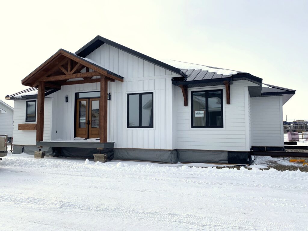 A small, modern white house with dark windows and a wooden porch stands against a snowy landscape under a bright sky, exuding a cozy and inviting feel.