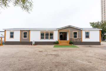 A modern, single-story manufactured home with a white exterior, brown trim, and gray stone accents. The front entrance features steps and greenery.