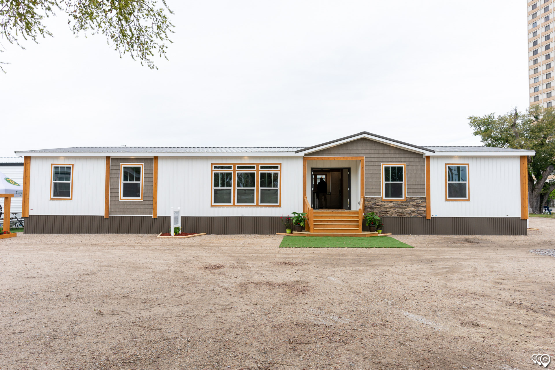 A modern, single-story manufactured home with a white exterior, brown trim, and gray stone accents. The front entrance features steps and greenery.