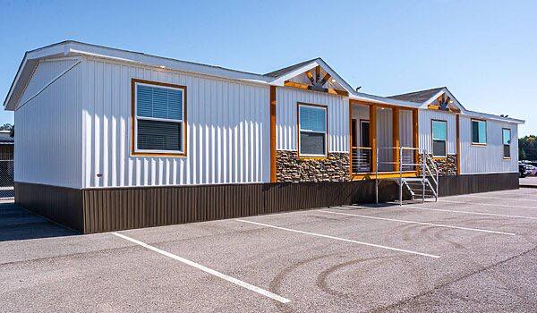 A modern mobile home with white paneled siding, stone accents, and several windows. It has a small porch with a ramp. The sky is clear and blue.