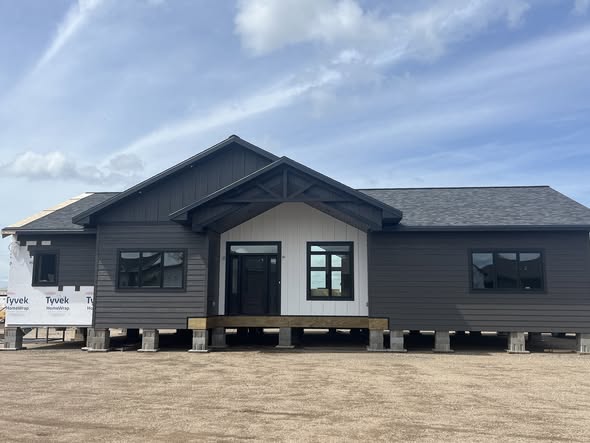 Single-story, dark gray house under construction, with white siding accents and black framed windows. Bright blue sky with soft clouds overhead.