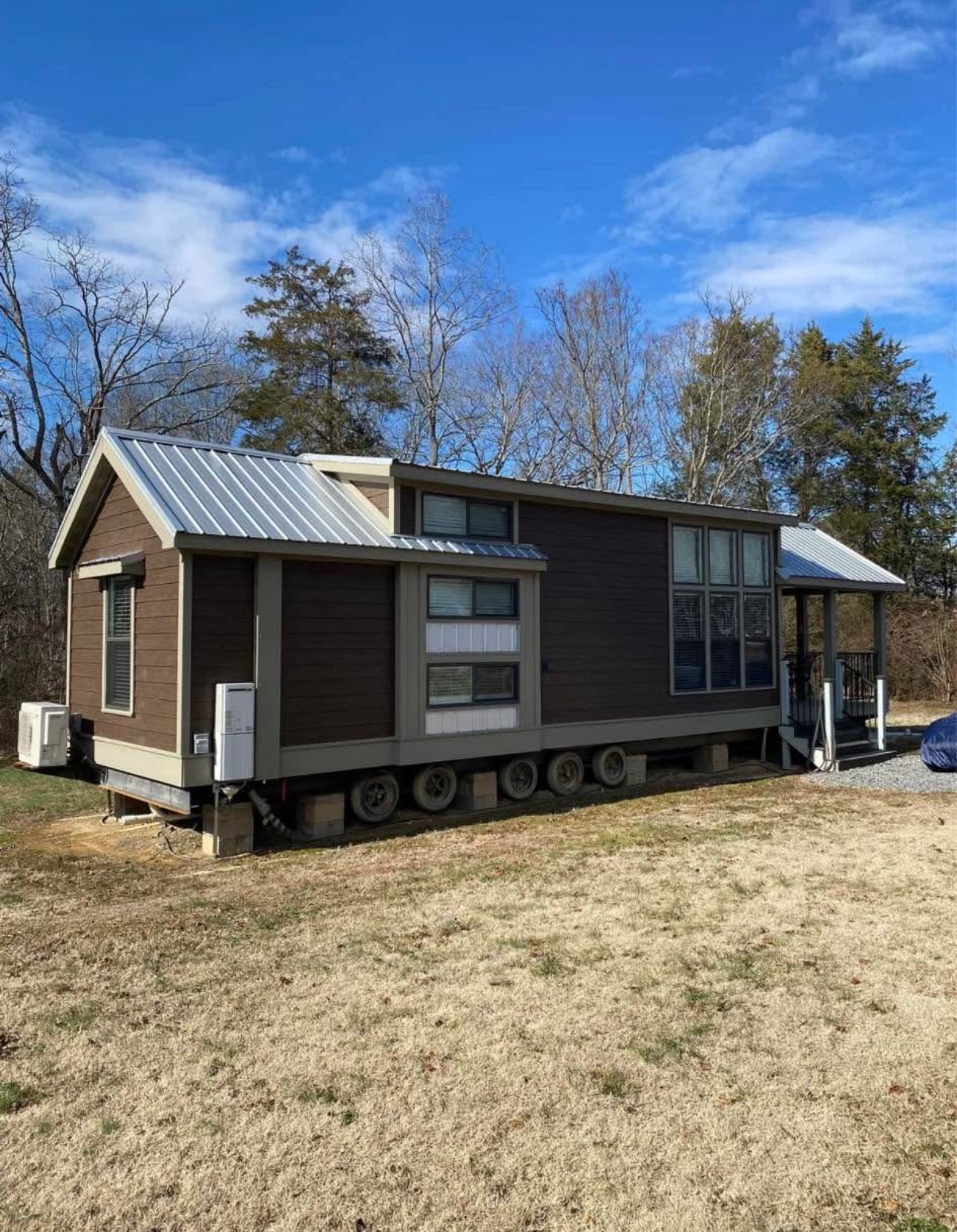 Small, mobile tiny house with brown wooden siding and a silver metal roof, set in a grassy area. Trees and a blue sky create a peaceful backdrop.