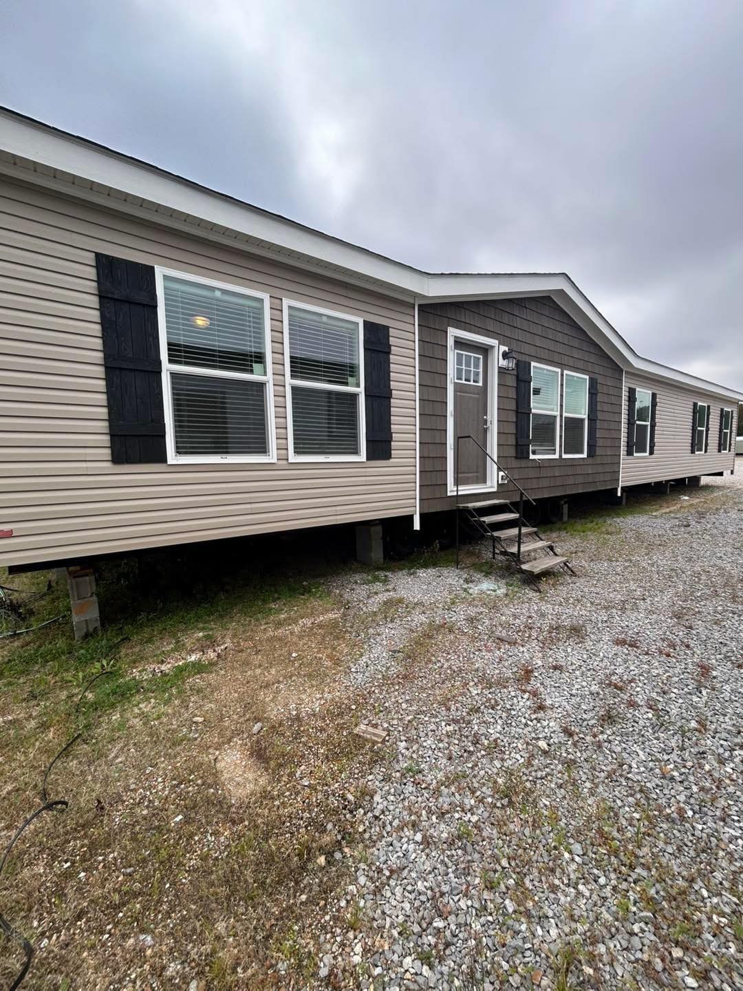 A beige manufactured home with black shutters, large windows, and a small metal staircase sits on a gravel and grass lot under a cloudy sky.