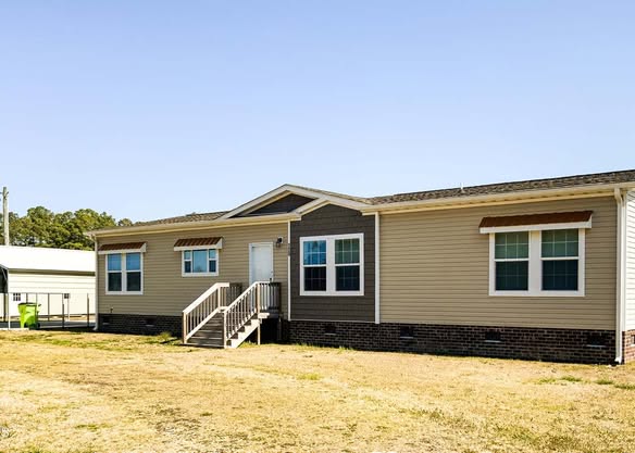 Single-story beige and brick mobile home with four windows, a white door, and small porch steps. It sits on a dry grass patch under a clear blue sky.
