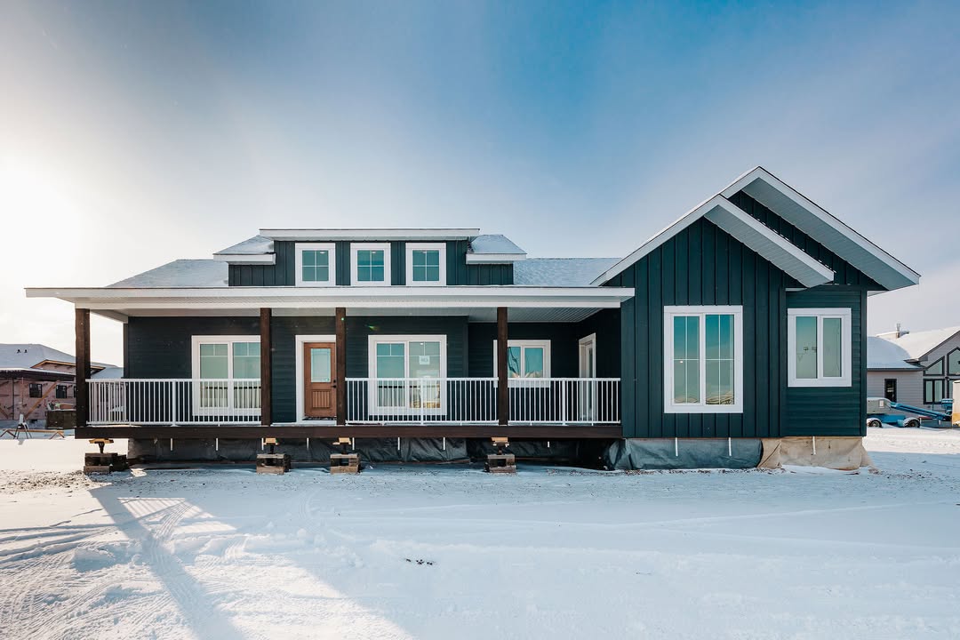 A modern, dark teal house with a white-trimmed porch stands elevated on a snowy landscape under a bright sky, conveying a sense of calm and elegance.