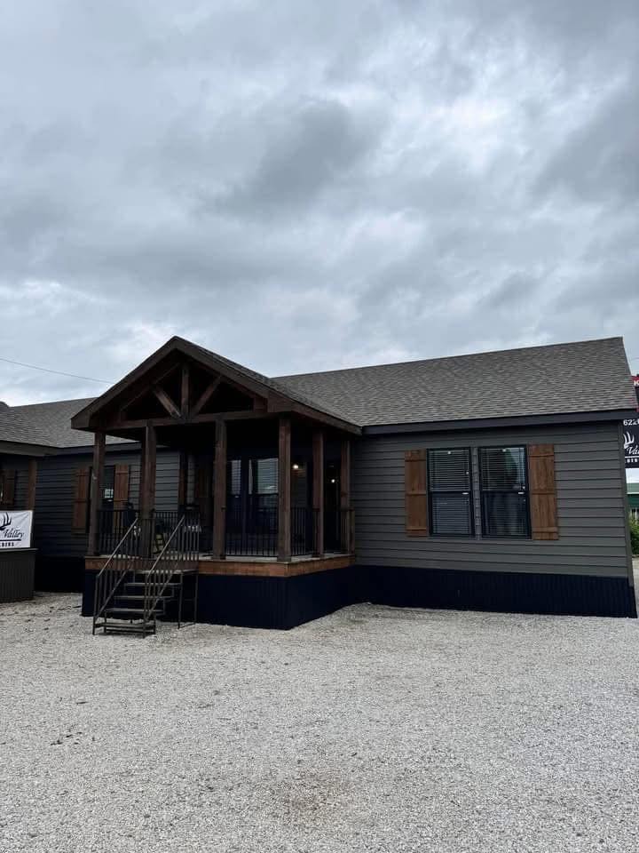 Single-story gray modular home with a raised wooden porch and dark wooden shutters. Cloudy sky overhead adds a somber tone. Gravel driveway in the foreground.