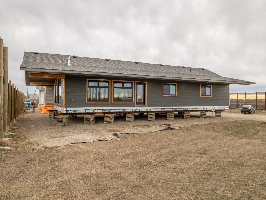 A modern gray home under construction, elevated on stacks of wooden pallets. The overcast sky sets a neutral tone. Bare ground surrounds the structure.