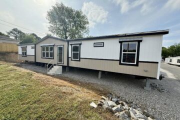 A beige and white mobile home with black trim sits on a gravel lot. Steps lead to the door. A tree stands behind, under a partly cloudy sky.