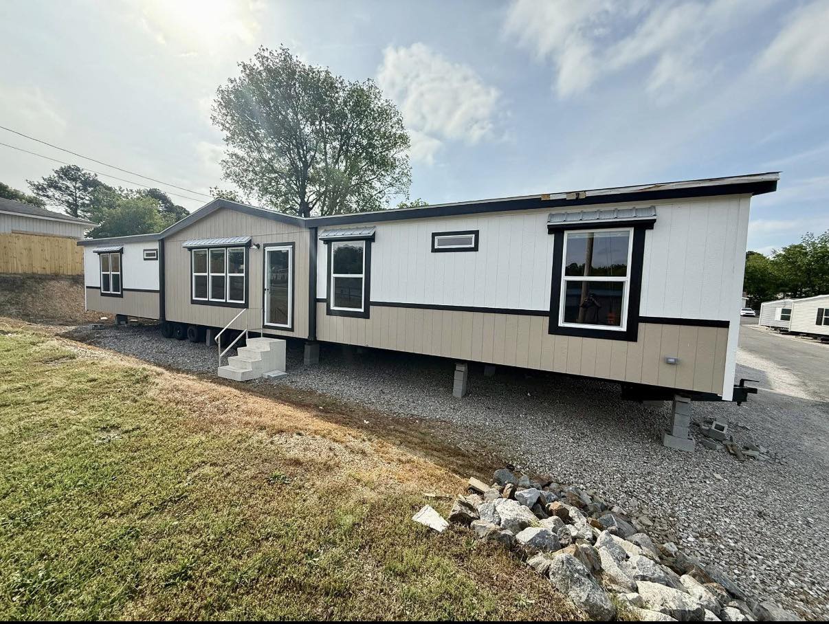 A beige and white mobile home with black trim sits on a gravel lot. Steps lead to the door. A tree stands behind, under a partly cloudy sky.