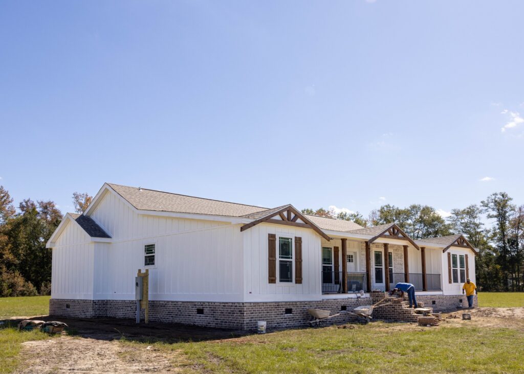 A white ranch-style house with brick foundation stands under a clear blue sky. Two people work on landscaping the front yard. The mood is tranquil and industrious.