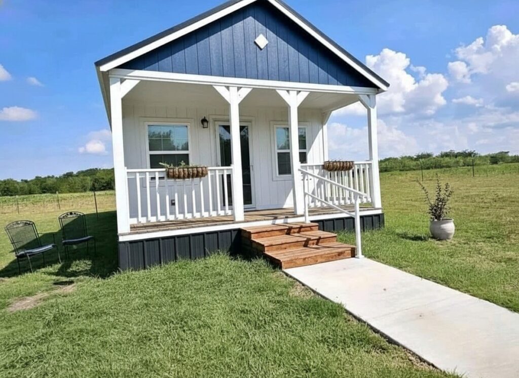 A small blue and white cottage with a porch sits on a grassy field under a bright blue sky. Two chairs are nearby, conveying a peaceful rural vibe.
