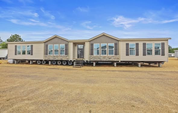 A beige manufactured home with multiple windows sits on a gravel lot under a clear blue sky, conveying simplicity and readiness for setup.
