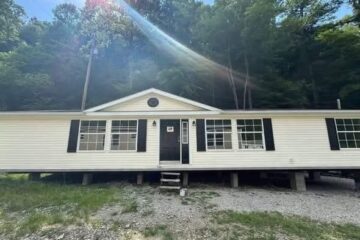 A single-story, white mobile home with black shutters, elevated on a gravel lot. Sunlight filters through trees in the background, creating a serene feel.