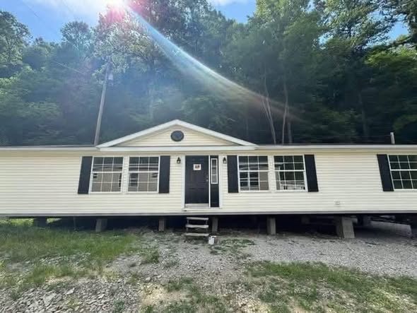 A single-story, white mobile home with black shutters, elevated on a gravel lot. Sunlight filters through trees in the background, creating a serene feel.