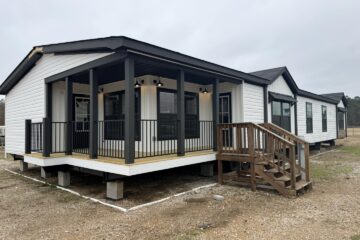Single-story modular home with white siding and black trim, featuring a covered porch with black railings and wooden steps. Overcast sky.