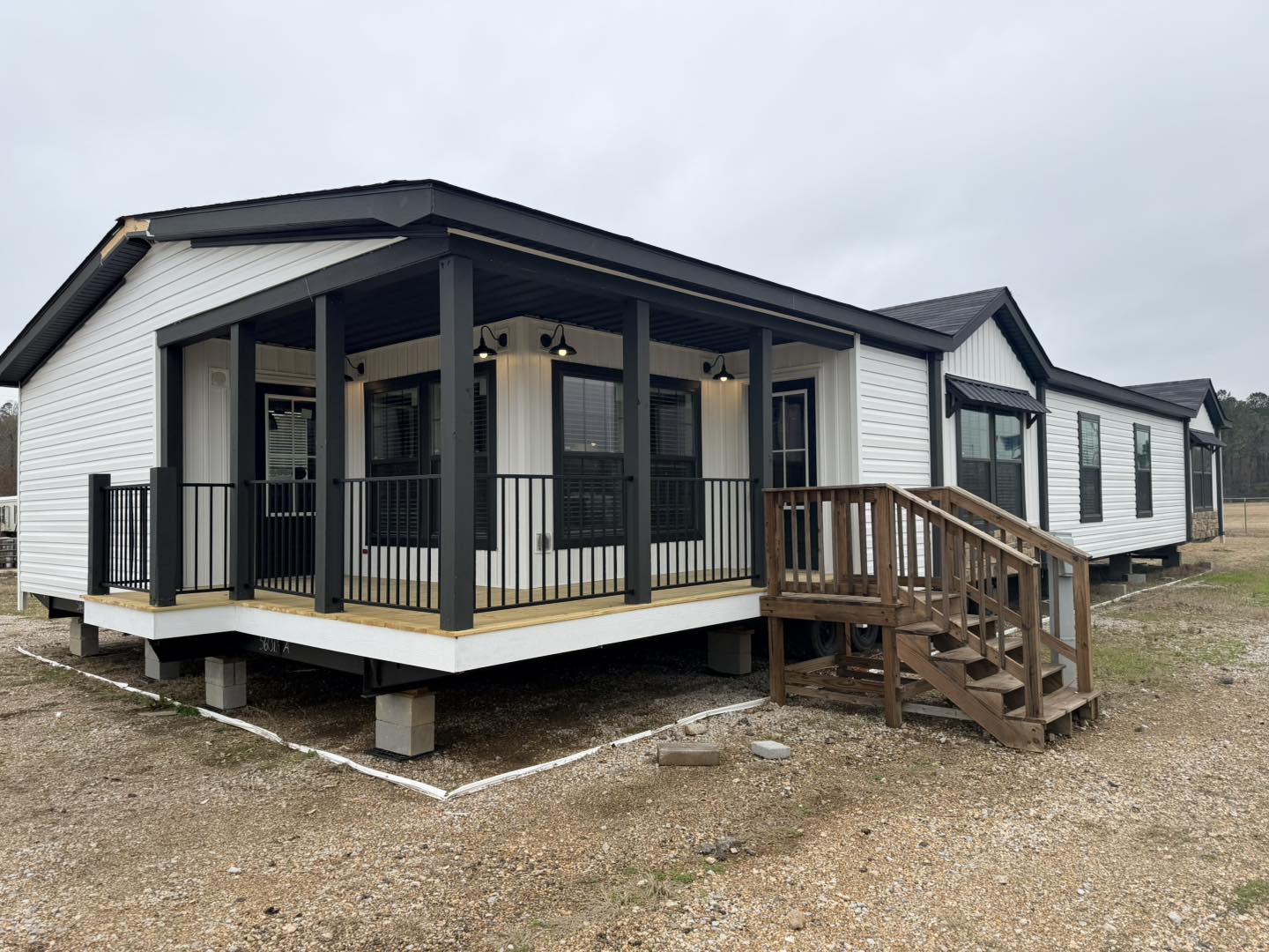 Single-story modular home with white siding and black trim, featuring a covered porch with black railings and wooden steps. Overcast sky.