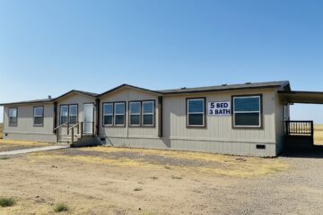 Single-story modular home with beige siding, labeled "5 Bed 3 Bath" in blue. It has multiple windows, a covered entrance, and is set on a dry, open landscape.