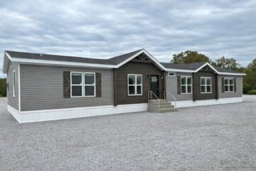 A modern, single-story modular home with brown siding, multiple white-framed windows, and a central front entrance with steps, set against a cloudy sky.
